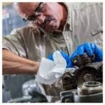 Una persona con gafas de seguridad y guantes azules se concentra en reparar maquinaria. Sostiene un paño blanco Wypall X70 mientras examina un componente metálico en un taller concurrido.