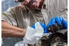 Una persona con gafas de seguridad y guantes azules se concentra en reparar maquinaria. Sostiene un paño blanco Wypall X70 mientras examina un componente metálico en un taller concurrido.